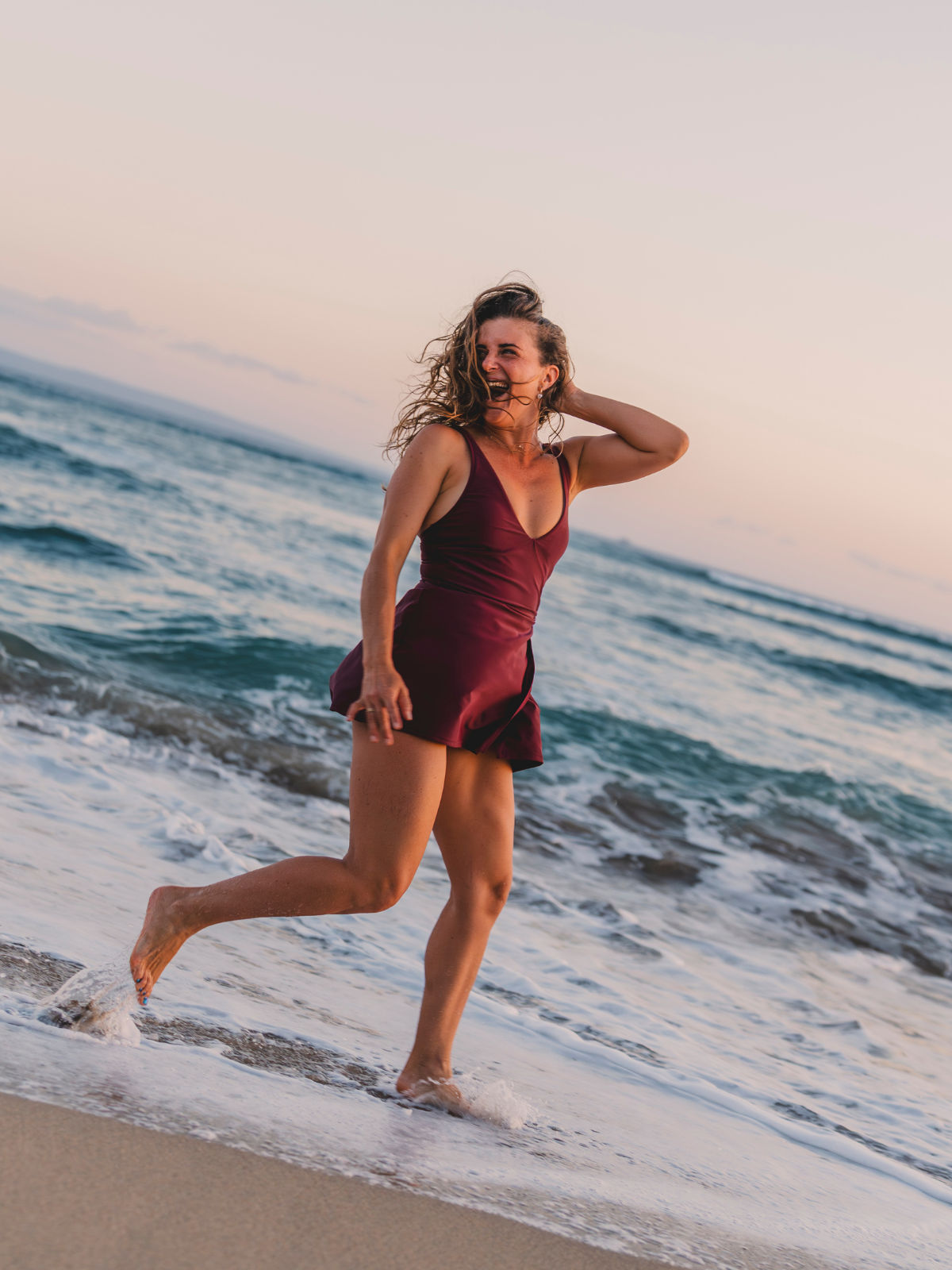 Woman joyfully walking along the beach wearing the Alana Swim Dress in a deep wine color, showcasing size-inclusive, supportive swimwear designed for every body with a comfortable fit.