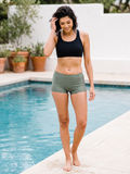 A woman wearing a black sports bra and olive green athletic shorts walks barefoot along the edge of a swimming pool.