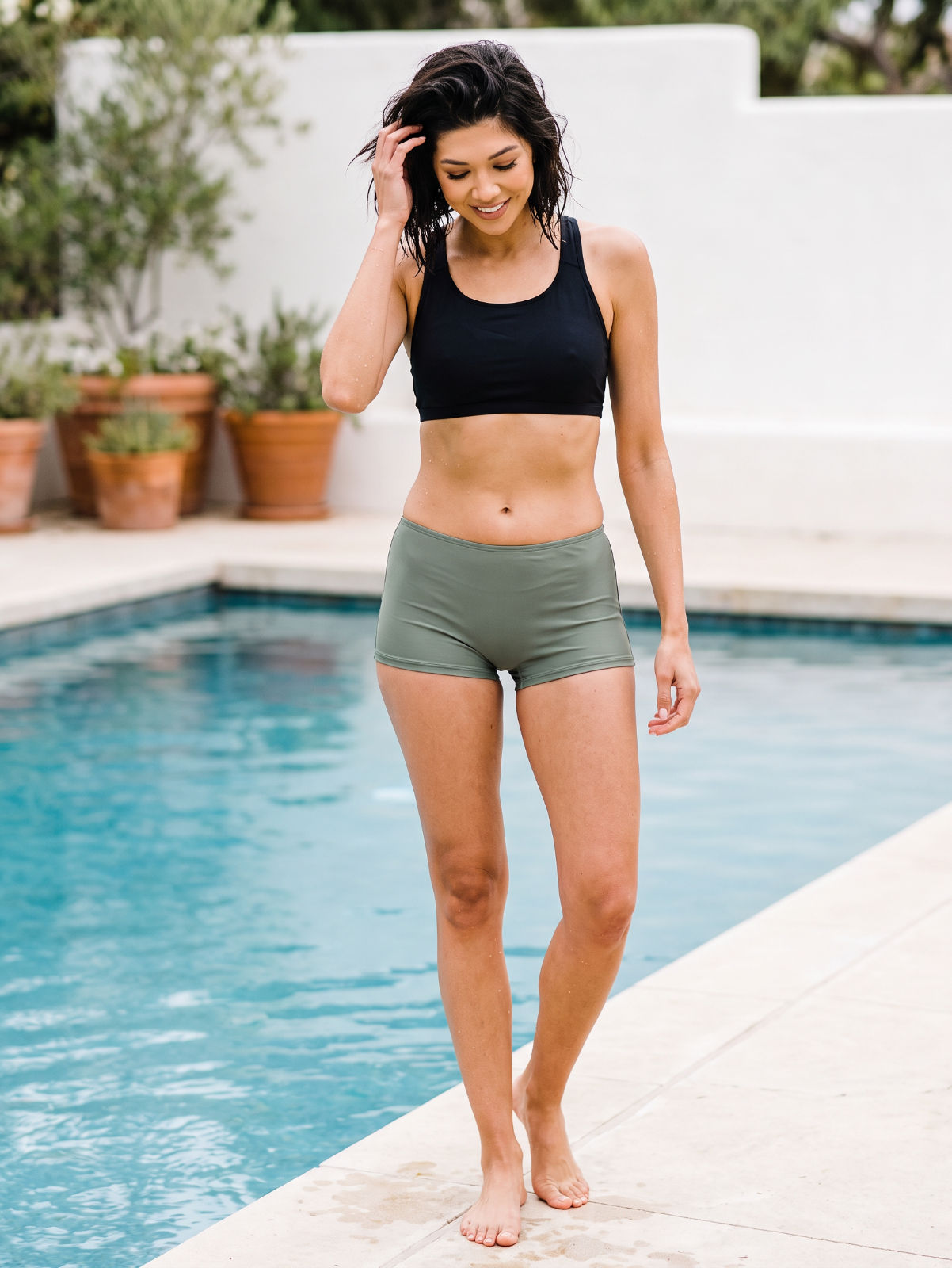 A woman wearing a black sports bra and olive green athletic shorts walks barefoot along the edge of a swimming pool.