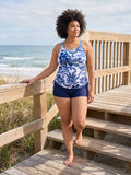 A woman with an afro hairstyle walks down wooden beach boardwalk stairs wearing a blue and white floral print tankini top and navy blue swim shorts, with the ocean and dunes in the background.