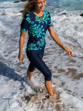 Woman wearing Capri Swim Leggings and a floral swim top walks through ocean waves showcasing modest supportive swimwear designed for every body