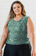 Woman wearing a green floral sleeveless top against a white brick wall.