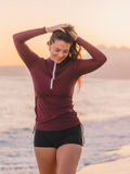 Woman wearing a maroon Half-Zip Nora Swim Top with long sleeves and black swim shorts at sunset on the beach, showcasing size-inclusive, supportive swimwear designed for every body.