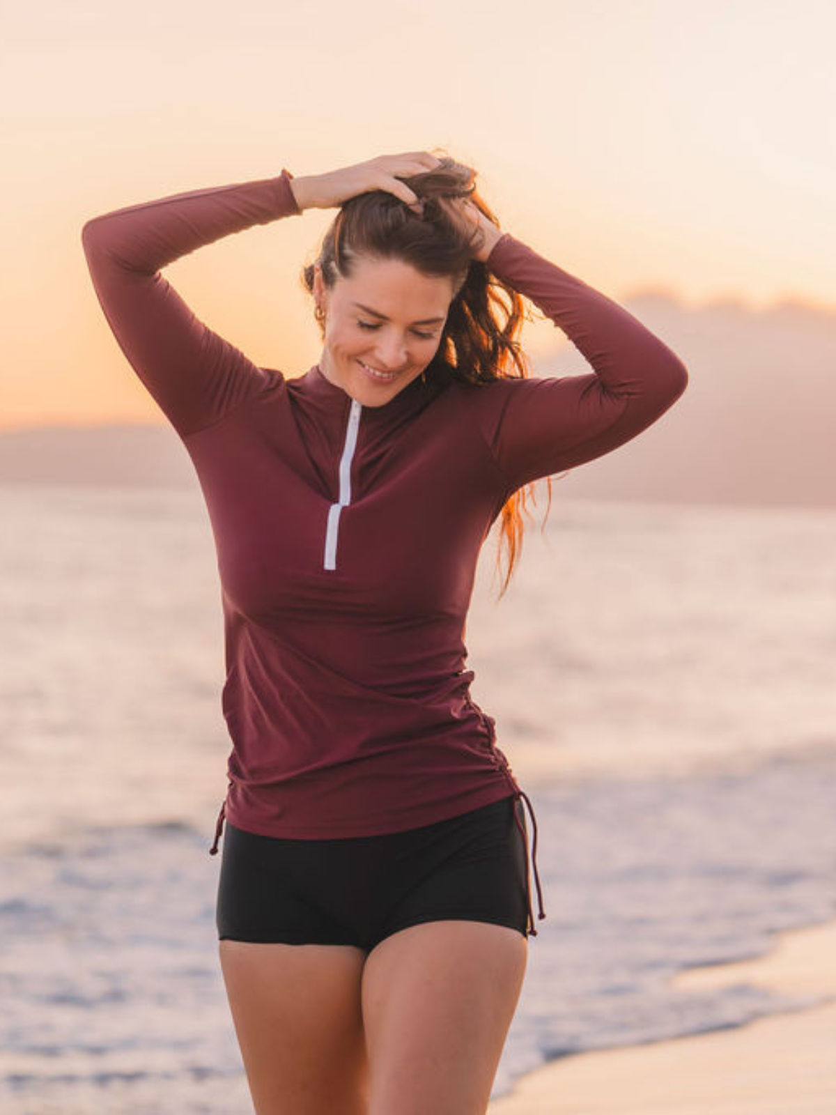 Woman wearing a maroon Half-Zip Nora Swim Top with long sleeves and black swim shorts at sunset on the beach, showcasing size-inclusive, supportive swimwear designed for every body.