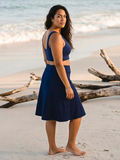 Woman wearing the Long Swim Skort With Capri Leggings in navy on a sandy beach with ocean waves behind her. Modest supportive swimwear designed for every body