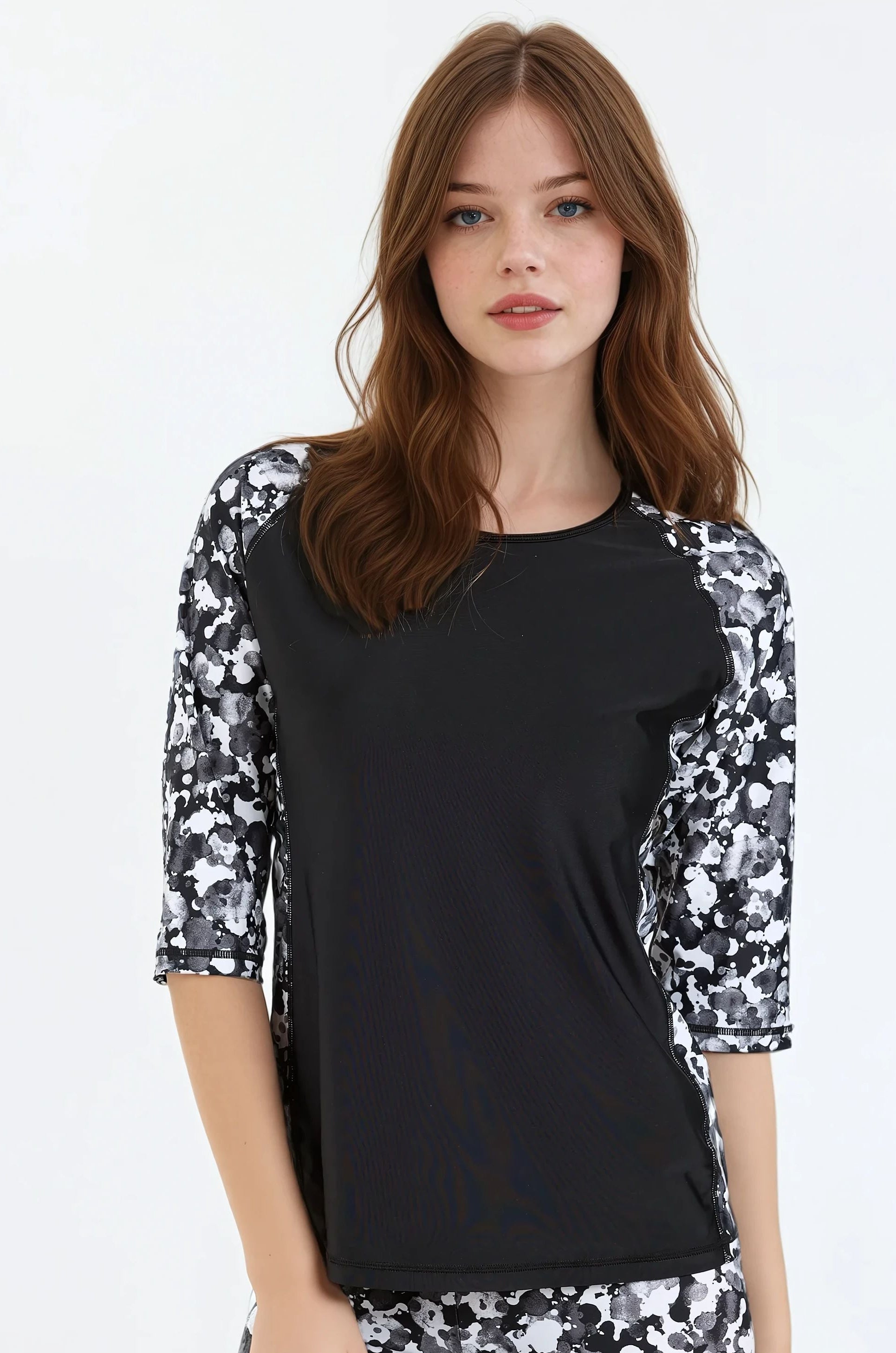 A model wearing a plus-size loose fit swim top with 3/4 length raglan sleeves and a round neckline. The top features green color with a floral pattern and contrasting side panels.