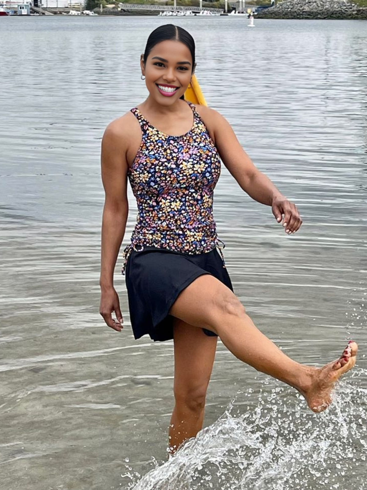 Woman wearing Maya Tankini Swim Top With Removable Cups | Final Sale in a colorful floral print with a black swim skirt, smiling and splashing water at the beach, showcasing size-inclusive, modest, supportive swimwear designed for every body.