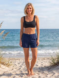 Sienna Swim Bra in black worn with navy swim shorts by a woman on a sandy beach showcasing supportive comfortable swimwear for every body