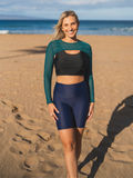 Woman standing on sandy beach wearing Tummy Control Long Bike Swim Shorts in navy, paired with a black and teal supportive swim top, showcasing size-inclusive, modest swimwear designed for every body.