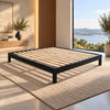 A modern black solid vic ash platform bed frame with natural wood slats, shown on a woven beige rug in a sunlit contemporary bedroom with a large window overlooking water.