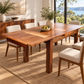 A rectangular wooden dining table with a warm medium-brown finish and matching upholstered chairs, styled in a bright modern dining room with large windows, plants, and a view of the water.