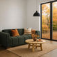 A modern living room scene featuring a dark green fabric sofa with orange and beige throw pillows, a round wooden coffee table, a textured beige rug, and a large black-framed glass door looking out onto an autumn forest.