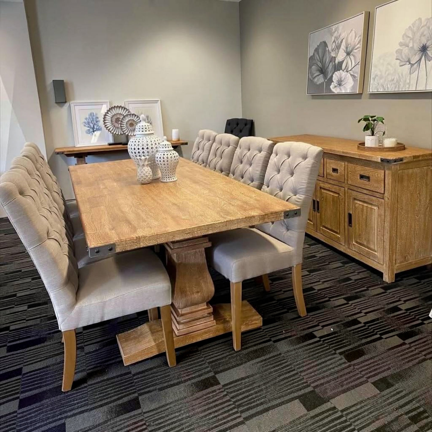 A wooden dining table with six tufted upholstered chairs in a staged dining room setting, featuring a matching wooden sideboard and patterned carpet.