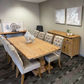 A modern dining room setup featuring a rectangular light oak wood dining table surrounded by tufted light gray upholstered chairs, with a matching wooden sideboard against a gray wall.