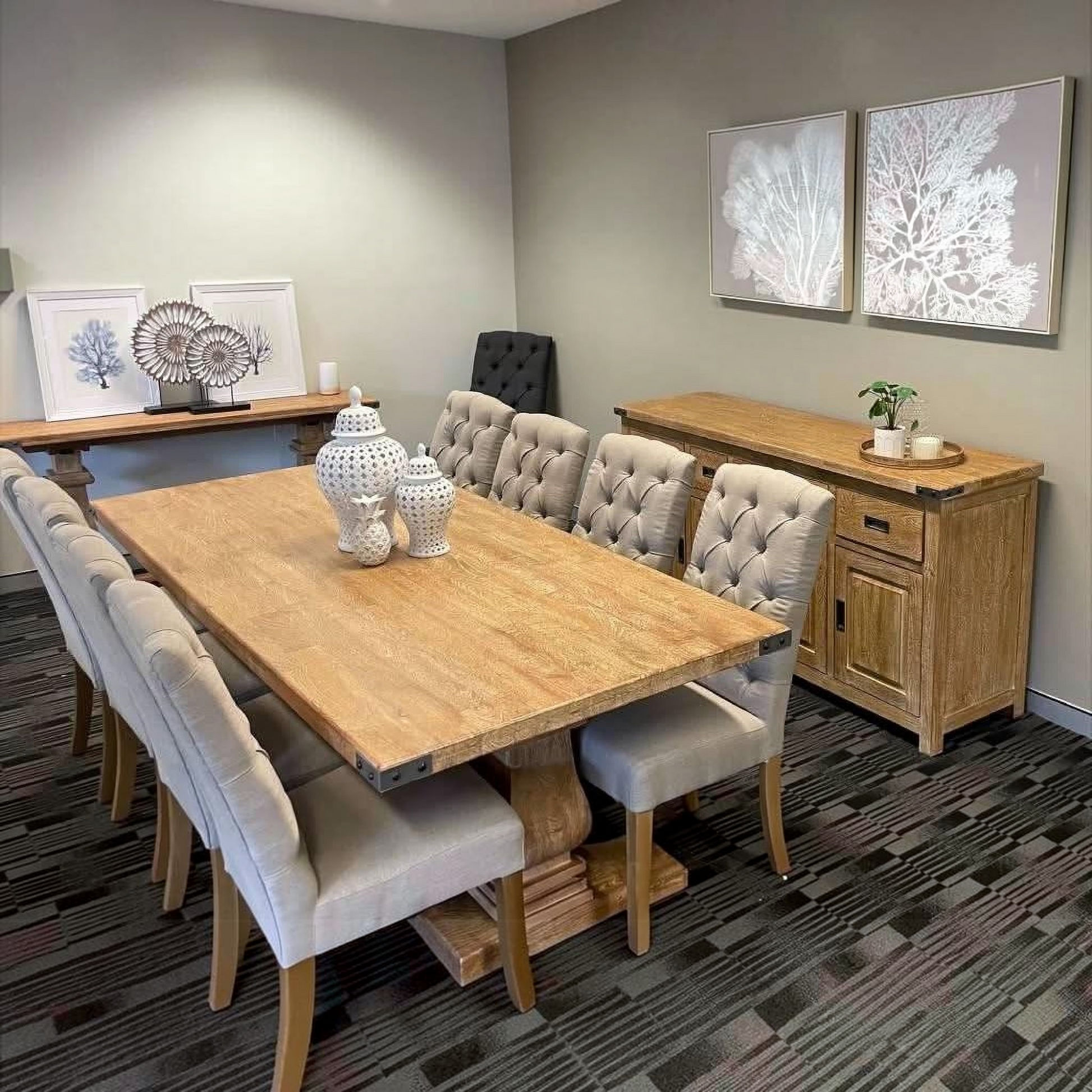 A modern dining room setup featuring a rectangular light oak wood dining table surrounded by tufted light gray upholstered chairs, with a matching wooden sideboard against a gray wall.