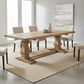 A rectangular wooden dining table with turned pedestal legs and a stretcher bar, shown in a bright dining room with four upholstered chairs, a rug, and wall art.