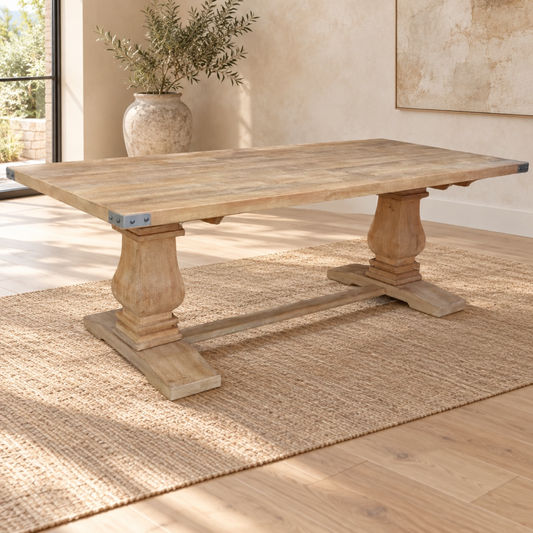 A rustic rectangular wooden dining table with turned pedestal legs and a stretcher base, shown in a sunlit room on a woven rug with a vase and framed wall art in the background.