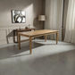 A rectangular wooden dining table with a natural oak finish is shown in a modern, sunlit room with gray concrete floors, white curtains, and a sideboard in the background.