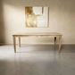 A light natural wood dining table with a rectangular top and four tapered legs, shown in a minimalist room setting with a concrete floor and a beige wall featuring abstract wall art.