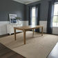 A rectangular wooden dining table with a natural oak finish stands on a large woven jute rug in a styled dining room with dark gray walls, white wainscoting, a white sideboard, and dark gray curtains.