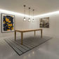 A modern dining room scene featuring a light oak rectangular table on a gray striped area rug, with three black pendant lights hanging above. The room has white walls, recessed lighting, and two framed abstract paintings.