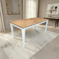 A rectangular dining table with a light oak wood top and white legs, placed on a textured cream rug in a bright, modern dining room.