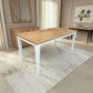A rectangular dining table with a light natural wood top and white legs, shown in a bright, modern living room setting on a textured light rug.