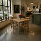 A rustic wooden dining table with four matching wooden chairs in a bright, modern cafe setting with a chalkboard menu, potted plants, and a green bar counter in the background.