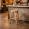 A light natural wood bar stool with a round seat and four tapered legs, positioned in front of a concrete-topped bar counter in a cafe setting.