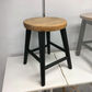 A rustic industrial-style stool with a round, weathered light brown wooden seat and four black legs, shown on a white table against a plain gray wall.
