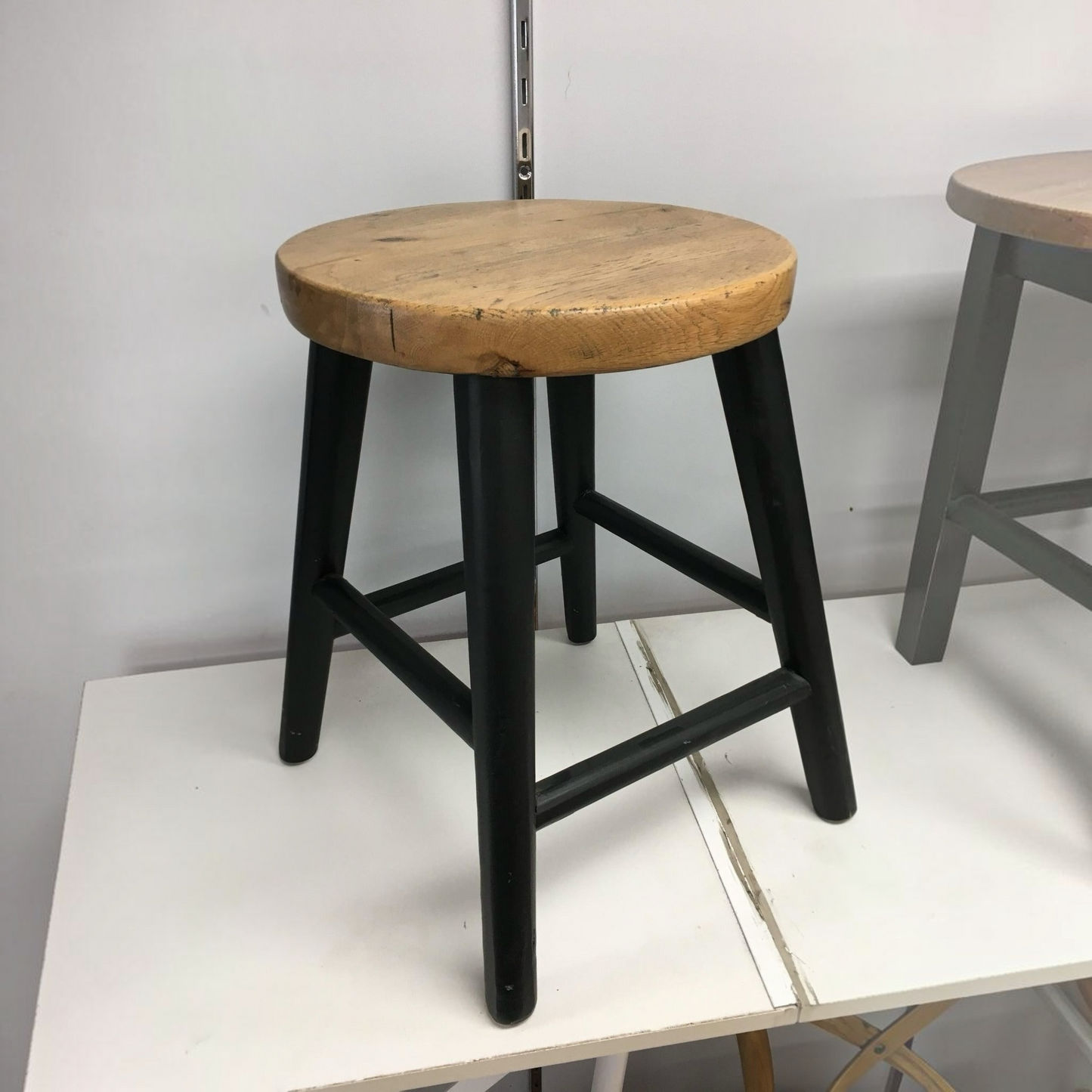 A rustic industrial-style stool with a round, weathered light brown wooden seat and four black legs, shown on a white table against a plain gray wall.