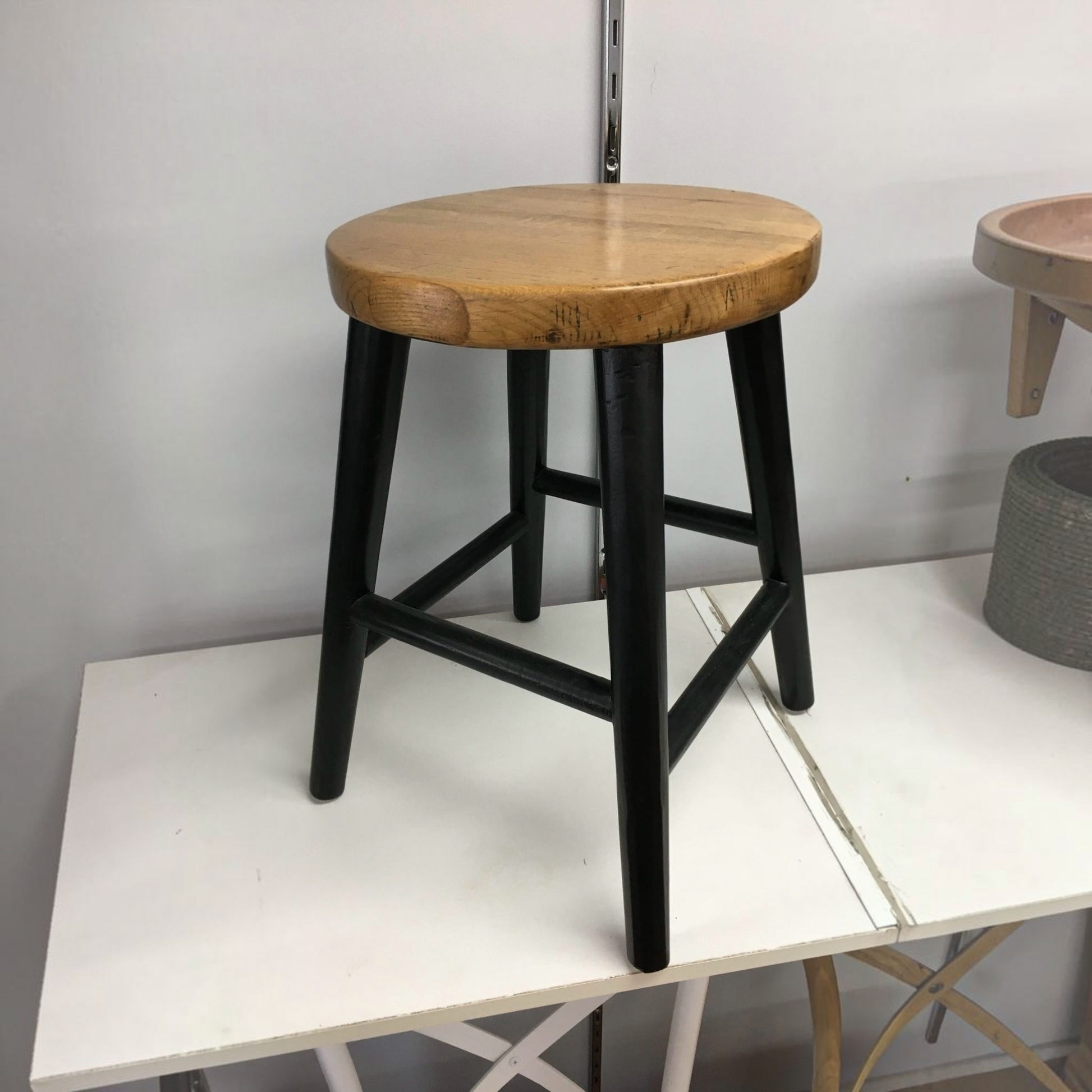 A rustic bar stool with a round natural wood seat and black metal legs, positioned on a white table against a light gray wall.