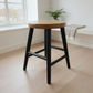 A modern bar stool with a round natural wood seat and black metal legs, positioned on a light wood plank floor in a bright, minimalist room.