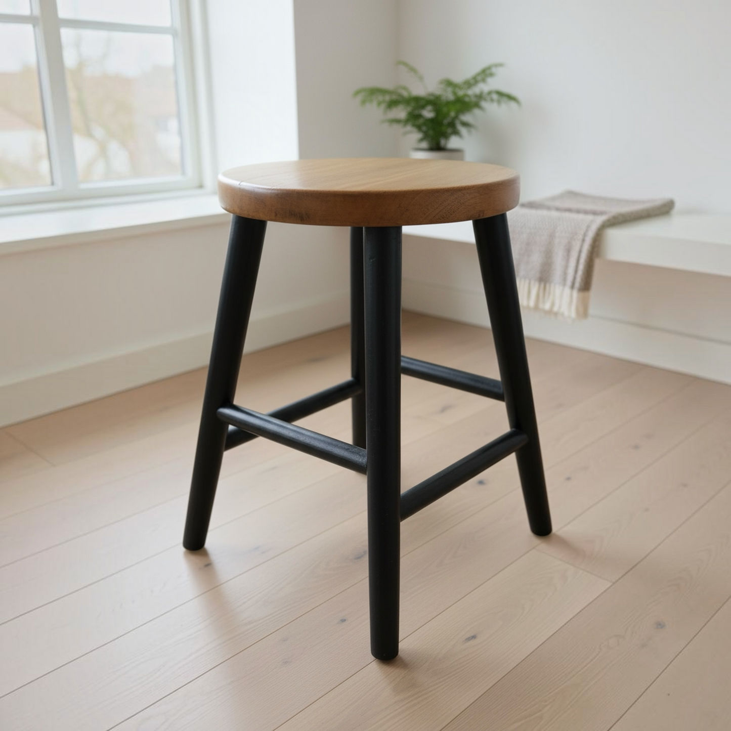 A modern bar stool with a round natural wood seat and black metal legs, positioned on a light wood plank floor in a bright, minimalist room.