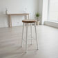 A modern bar stool with a round natural wood seat and four painted white legs, positioned in a bright, minimalist room with light wood floors and a console table in the background.