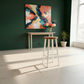 A modern bar stool with a light wood seat and white legs stands in a bright, minimalist room with a dark green wall, a console table, and a colorful abstract painting.