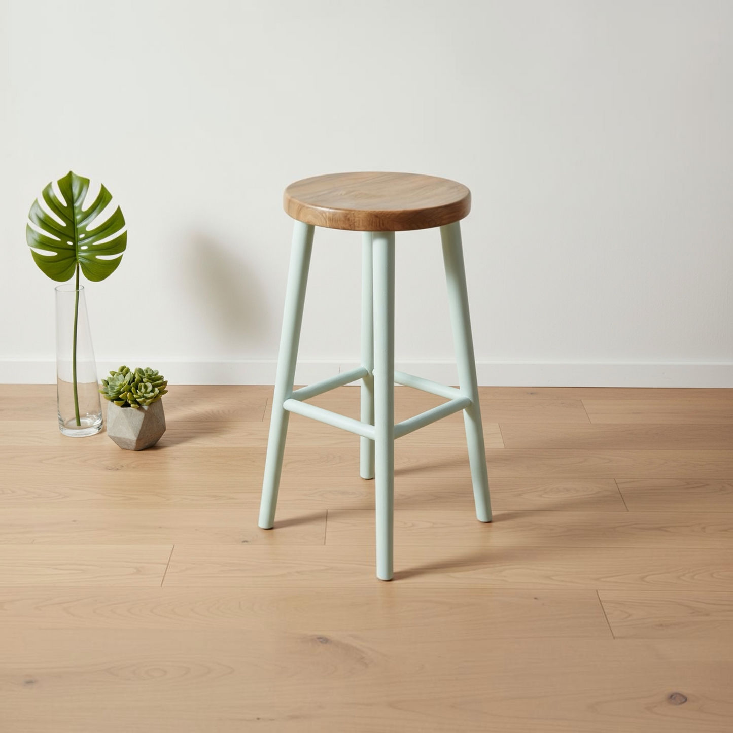 A mint green bar stool with a round natural wood seat and a light wood floor, styled in a minimalist room with a white wall and small plants in a glass vase.