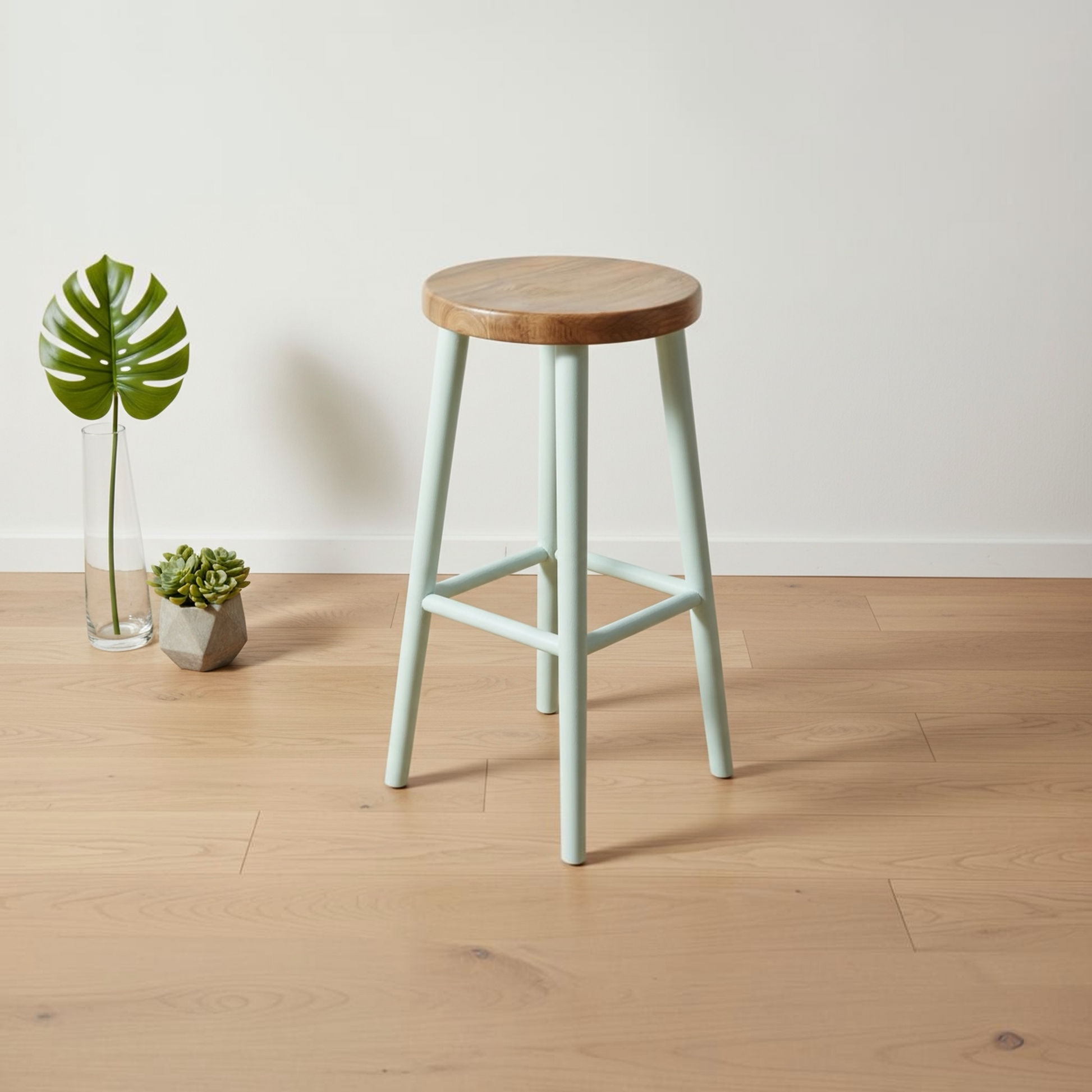 A mint green bar stool with a round natural wood seat and a light wood floor, styled in a minimalist room with a white wall and small plants in a glass vase.