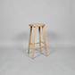 A light natural wood bar stool with a round seat and four tapered legs, shown on a neutral gray studio background.