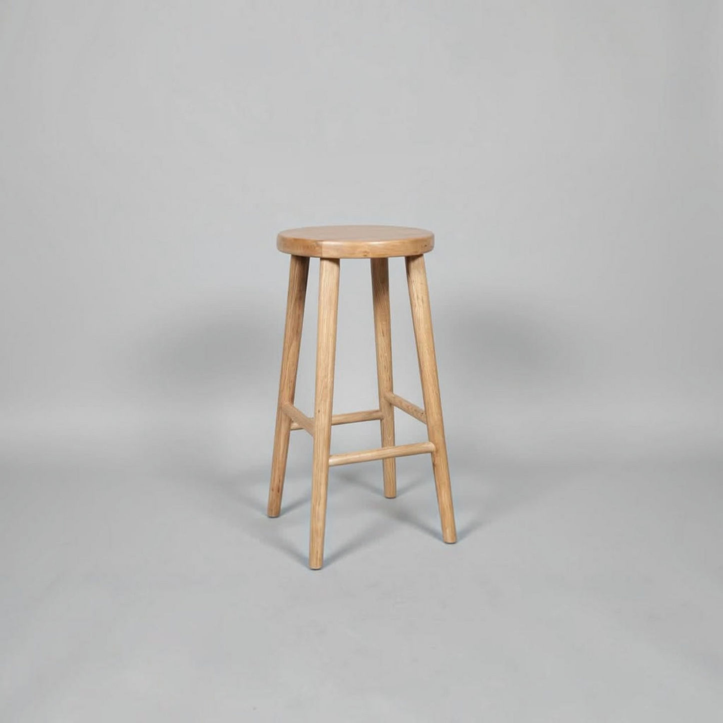 A light natural wood bar stool with a round seat and four tapered legs, shown on a neutral gray studio background.