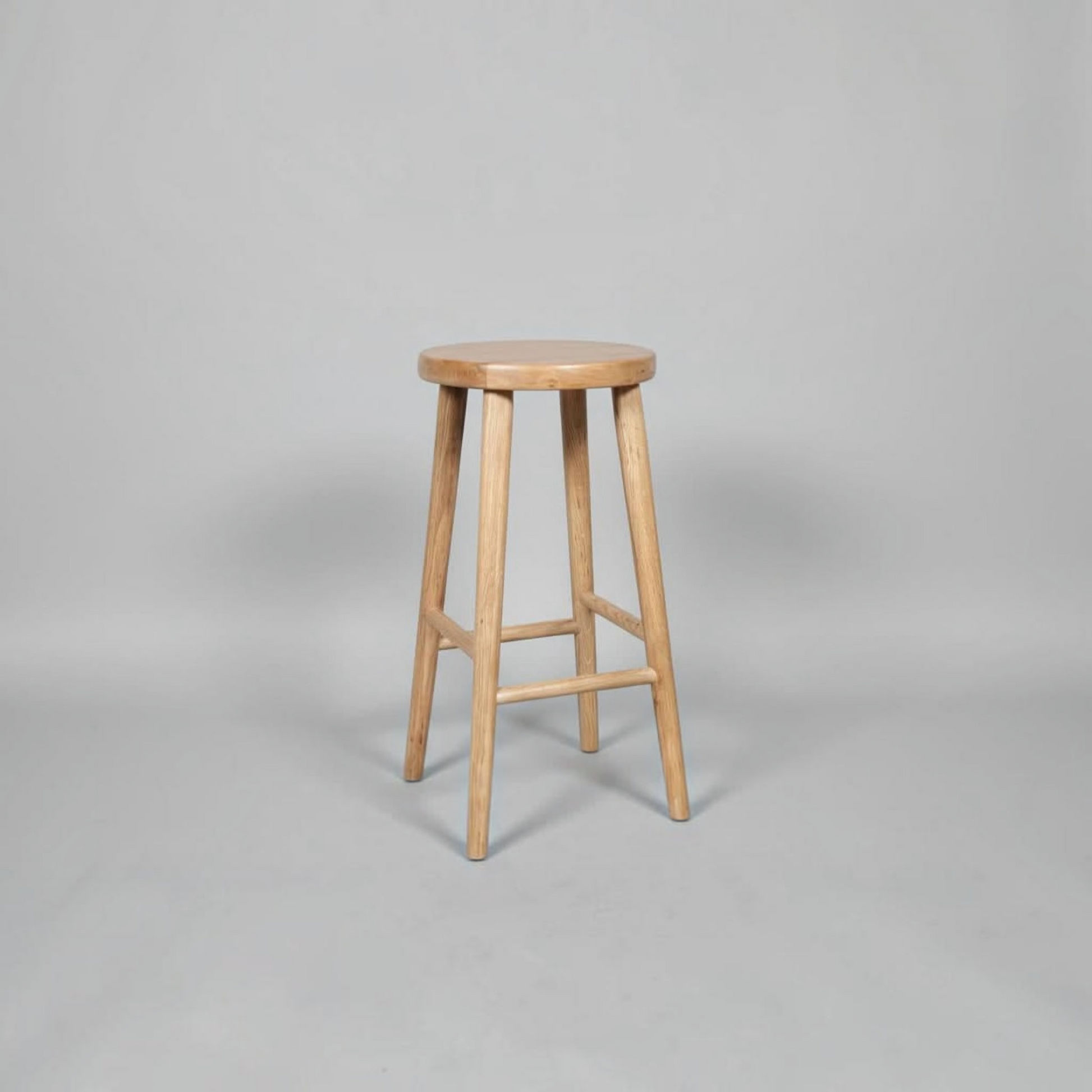 A light natural wood bar stool with a round seat and four tapered legs, shown on a neutral gray studio background.