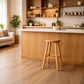 A light natural wood bar stool with a round seat and four legs stands in front of a modern kitchen island with a white countertop and warm wood cabinetry.