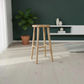 A light natural wood bar stool with a round seat and four legs, shown in a modern room setting with a dark green wall, white floating shelf, and a potted plant.
