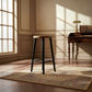A wooden bar stool with black legs stands on a patterned area rug in a sunlit room with a desk and window in the background.