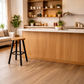 A modern kitchen island with a light wood base and white countertop, featuring a round wooden bar stool with black legs in the foreground. The room has light wood flooring, white subway tile backsplash, open wooden shelves with decorative items, and potted plants.
