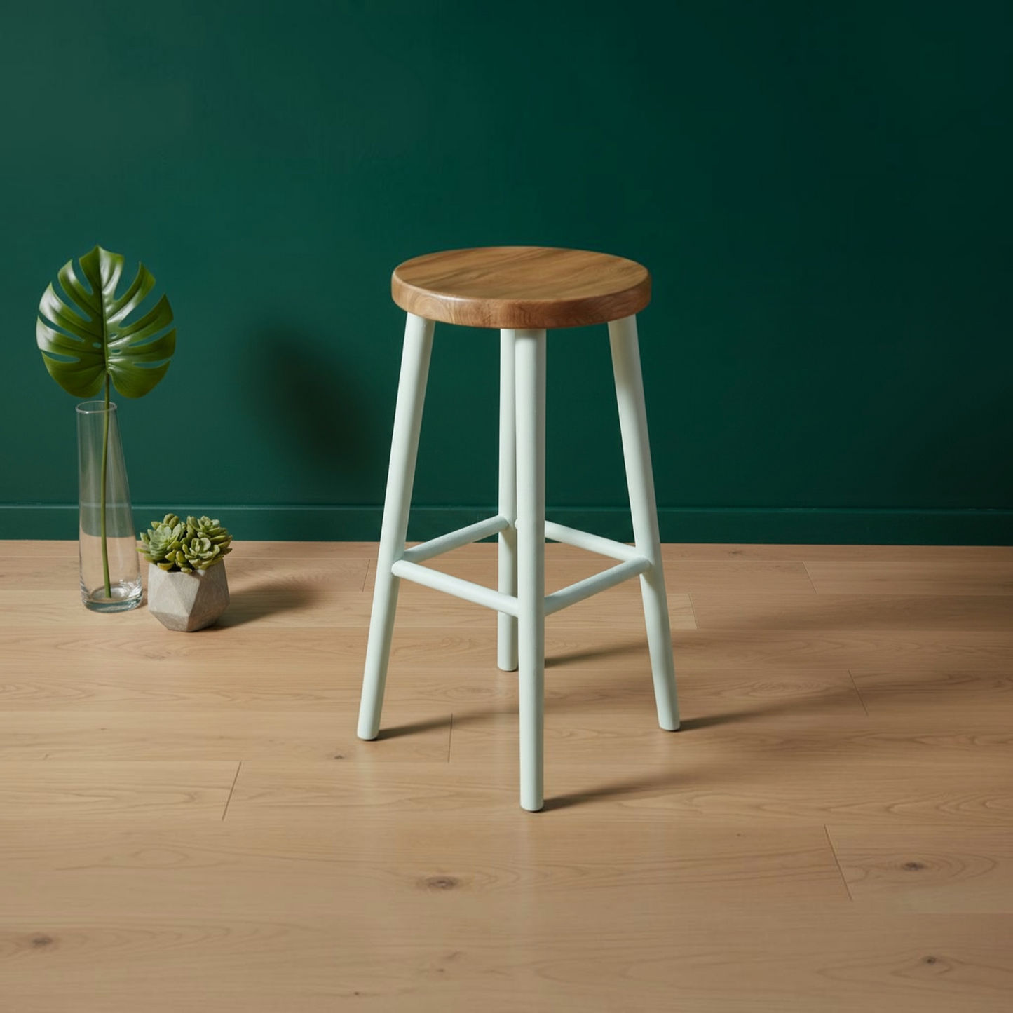 A modern bar stool with a round natural wood seat and pale mint green legs, set on a light wood floor against a dark green wall with small potted plants in the background.