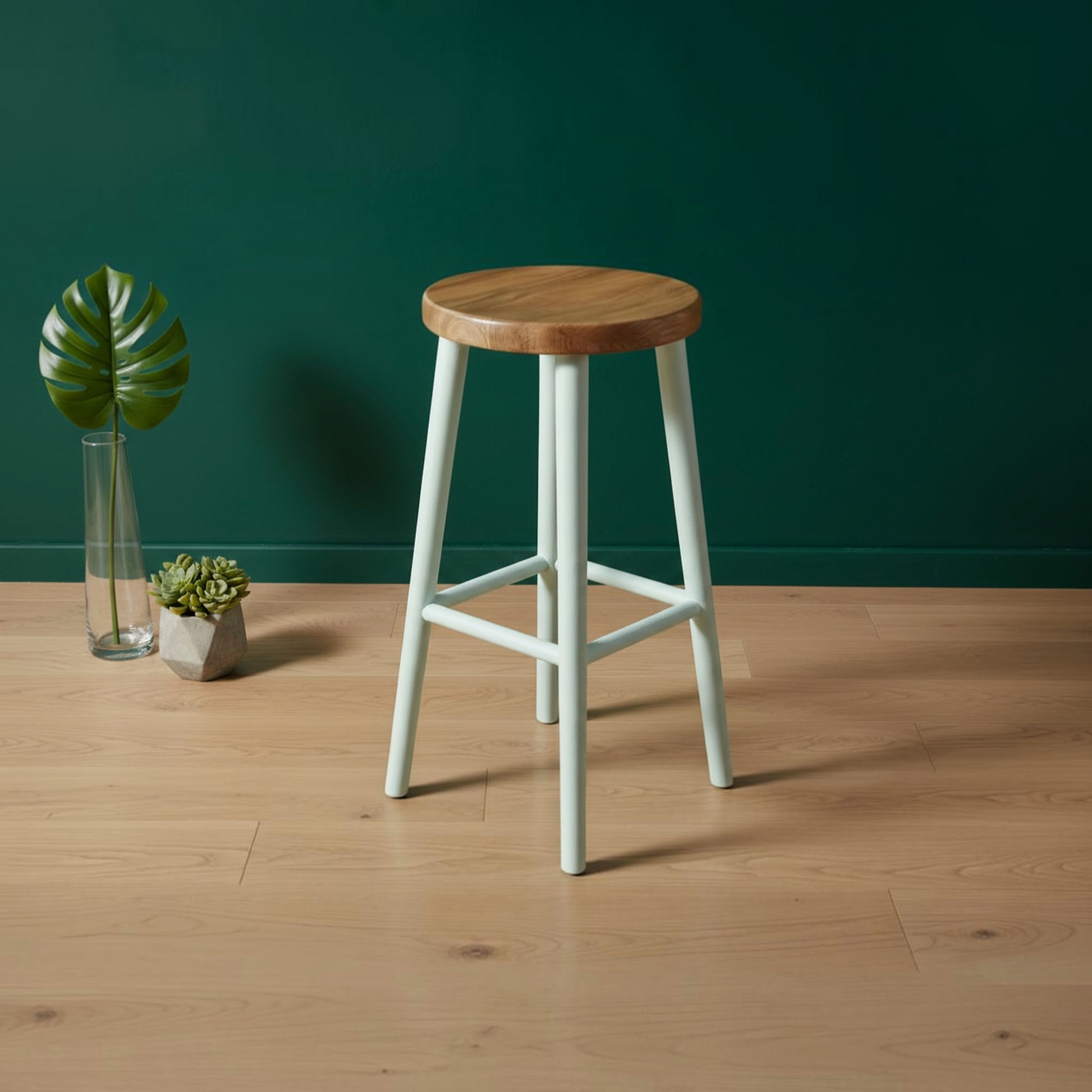 A modern bar stool with a round natural wood seat and pale mint green legs, set on a light wood floor against a dark green wall with small potted plants in the background.