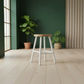 A modern bar stool with a light natural wood seat and white legs, positioned in a minimalist room with a dark green accent wall, light wood flooring, and potted plants.