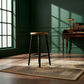 A wooden bar stool with a light oak seat and black legs stands on a vintage-style patterned rug in a sunlit room with dark green paneled walls and a desk.