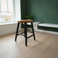 A modern bar stool with a round natural wood seat and black metal legs, positioned in a bright room with light wood floors, a dark green accent wall, and a built-in bench.
